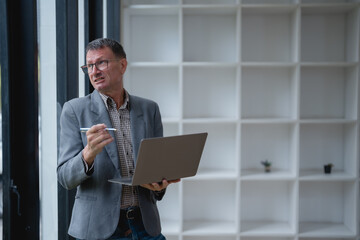 Senior manager explaining concepts while holding a laptop and pen, standing confidently in a modern office environment, embodying business communication and collaboration