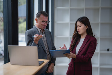 Senior male manager explaining marketing strategy using laptop to young businesswoman holding clipboard in modern office, she seems confused and worried about new project