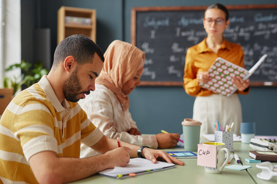 Young adult Caucasian man and young adult Muslim woman wearing hijab writing in notebooks during language school lesson, female teacher standing in background holding book near chalkboard - Powered by Adobe