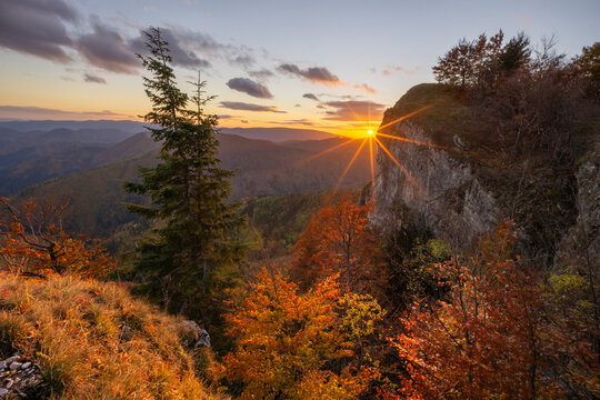 View of the sun bursting over the cliffside, casting a warm glow on the autumn trees and distant mountains, Majerova skala rock, StarÃ© Hory, BanskobystrickÃ½ kraj, Slovakia.