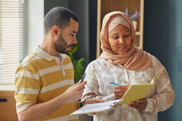 Young adult Middle Eastern man and woman discussing notes and holding pens and notebooks in language school classroom, collaborating on educational activity