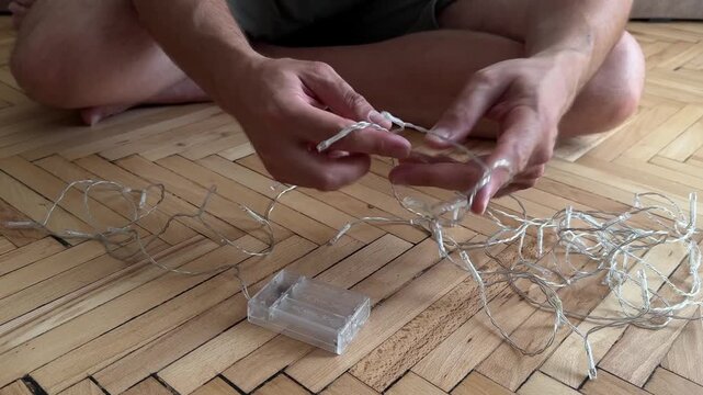 Close-up of a man's hands untangling a string of small LED fairy lights on a wooden floor. Represents home decor, DIY projects, and holiday preparation