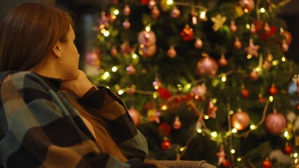 Woman Sitting Indoors Looking at Decorated Christmas Tree with Warm Lights in Soft Focus Close Angle, Holiday Reflection Concept