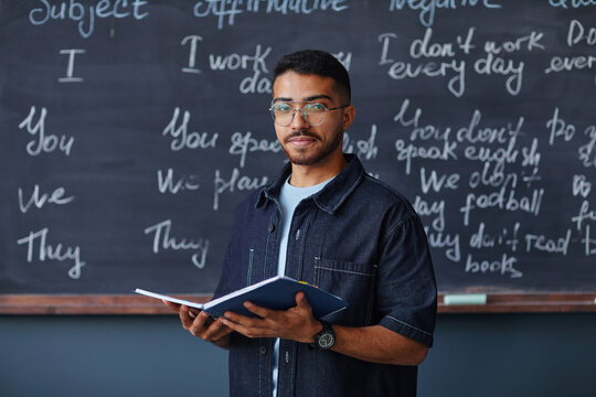 Portrait of young adult Middle Eastern man standing in front of chalkboard holding open notebook, wearing glasses, looking into camera, teaching language lesson in classroom setting - Powered by Adobe