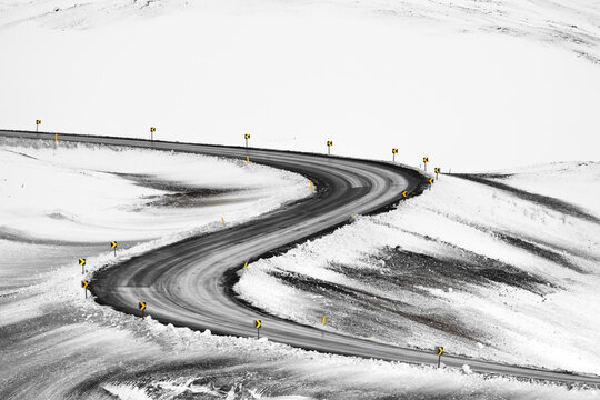 View of a winding road cuts a dark swathe through the stark white snow-covered landscape, punctuated by warning signs, in Iceland, Iceland.