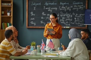 Young adult Caucasian woman holding digital tablet standing in front of multiethnic group of young adults sitting at table with British and American flags, studying English language in classroom