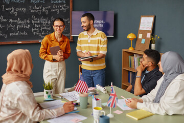 Diverse group of young adults participating in language school lesson, two instructors standing and smiling while students sitting at table with notebooks and flags © Seventyfour
