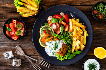 Fried salmon steak with tzatziki sauce, homemade potato fries and tomato salad on wooden table