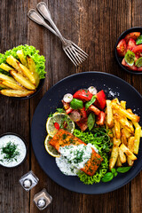 Fried salmon steak with tzatziki sauce, homemade potato fries and tomato salad on wooden table