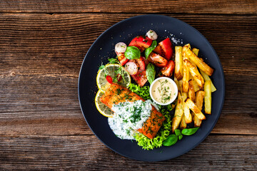Fried salmon steak with tzatziki sauce, homemade potato fries and tomato salad on wooden table