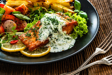 Fried salmon steak with tzatziki sauce, homemade potato fries and tomato salad on wooden table