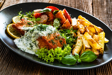 Fried salmon steak with tzatziki sauce, homemade potato fries and tomato salad on wooden table