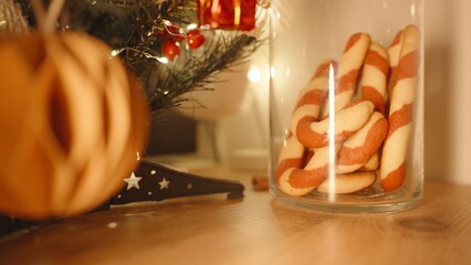 Candy Cane Cookies Displayed in Tall Glass Jar Near Decorated Tree on Wooden Table, Side Angle, Festive Cookie Display Concept