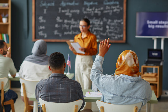 Diverse group of young adults and teenagers sitting in classroom raising hands, while female teacher standing in front of chalkboard holding papers during language school lesson
