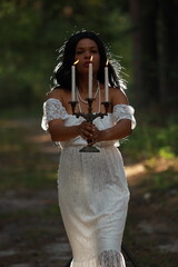 Naklejka premium Young curly dark-haired woman in white dress with vintage candlestick in evening forest