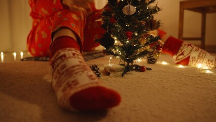 Woman in Red Pajamas Sitting Behind Mini Tree with One Foot in Festive Sock Extended Toward Camera, Low Angle, Cozy Holiday Vibes Concept
