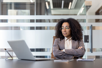 A serious young African American businesswoman is sitting at a desk in the office, crossing her arms over her chest and looking thoughtfully to the side