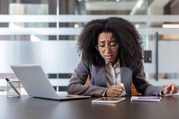 African American young businesswoman sitting sick at her desk, holding a napkin and suffering from pain