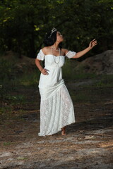 Fototapeta premium Young beautiful dark-haired curly woman in a white dress with a crown on her head in the forest