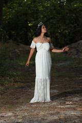Fototapeta premium Young beautiful dark-haired curly woman in a white dress with a crown on her head in the forest