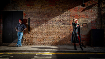 A Working Relationship. A sex worker stands waiting for business on a city pavement at night. Her guardian is in the background acting as protection. From an image series featuring character models.