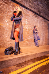 A Working Relationship. A sex worker stands waiting for business on a city pavement at night. Her guardian is in the background acting as protection. From an image series featuring character models.