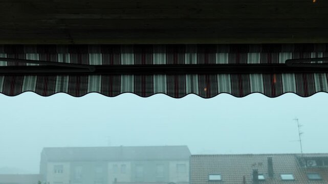 Heavy rain falling on a red and white striped awning in a gloomy urban landscape, with distant buildings obscured by fog on a dark and stormy day