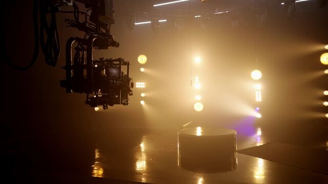 Professional camera and stabilizer rig hanging on a crane in a dark and foggy studio, with bright golden spotlights illuminating the empty stage, conveying cinematic production