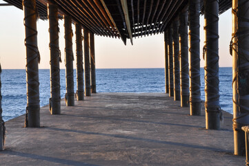 Fototapeta premium Symmetrical view under concrete pier with rope-wrapped columns facing the sea at sunset, architectural perspective and marine atmosphere, scenic seascape and travel destination