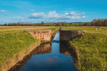 Fototapeta premium A simple water gate manages flow in a dry channel beside a grassy field in a marsh area under clear skies with blue reflections in the water.