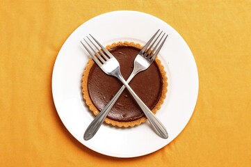 A rich chocolate tart served on a white dish with two crossed forks, set against a vibrant and playful tablecloth.