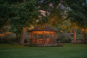 Obraz premium A charming pavilion glowing with fairy lights at twilight, nestled among verdant trees and manicured grass, exuding a welcoming and comfortable vibe.