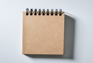 Top-down, blank, brown, square, spiral-bound notebook casting a shadow on a bright, white surface