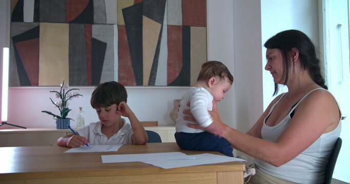 Boy working on homeschooling assignments at the table while mother cradles baby, demonstrating concentration and family-oriented homeschooling in a peaceful home setting focused on education