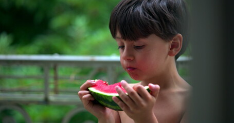 Boy deeply focused on eating a slice of watermelon, enjoying the juicy fruit outdoors on a balcony, savoring the moment in a peaceful and relaxed setting