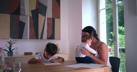 Tired boy rubbing his eye while doing homeschooling work at the table, mother holding baby nearby...