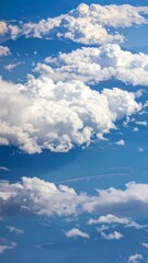 A vast expanse of brilliant blue sky, dotted with fluffy, white cumulus clouds, reveals a mountain range below, providing a serene and peaceful vista from an elevated perspective.