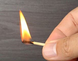 A lit match held by a hand against a gray surface, showcasing the flame and the wooden matchstick.