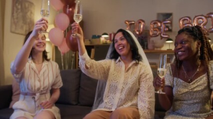 Lady of hour with veil clinking glasses full of champagne with her happy biracial friends while they sitting in living room fully prepared for bachelorette party
