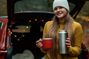 Cute teenage girl drinks hot tea from a large thermos in the trunk of a red car in an autumn forest. Picnic.