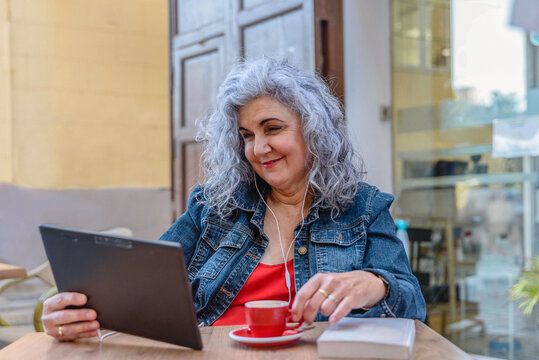 Smiling senior woman using earphones and digital tablet and drinking coffee at outdoor cafe