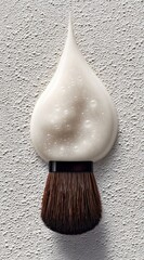 Teardrop of pale cream rests on a brown bristle brush against a textured off-white background