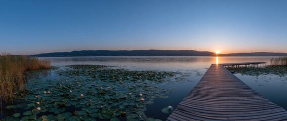 Panoramic lake scene, with lily pads in foreground, pier leading to sunset over distant mountains, beneath clear blue sky, serene landscape