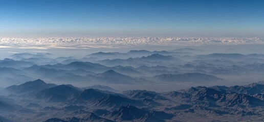 A scenic view of hazy, blue-toned mountains seen from high above, with a clear sky and a distant layer of fluffy clouds on the horizon
