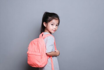 A young girl with dark hair, wearing a gray shirt and a salmon-colored backpack, looks over her shoulder against a solid gray backdrop