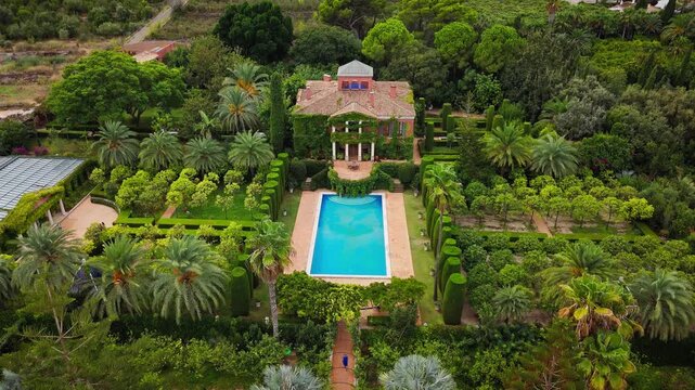 Drone video of a Spanish public garden featuring a historic ivy-covered building, elegant topiary, palm trees, and a pool surrounded by lush greenery.