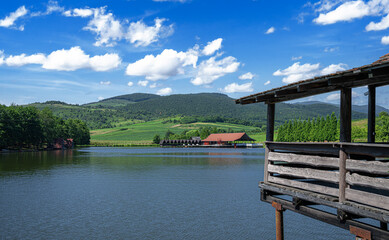 Fototapeta premium A serene lakeside view on a sunny day with green hills and a red-roofed building in the distance, framed by a rustic wooden deck in the foreground.