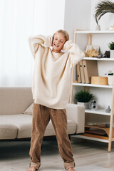 Young person smiles while stretching in a cozy living room, wearing a soft cream sweater and patterned pants, warm natural light, calm home scene, comfortable mood and ease.