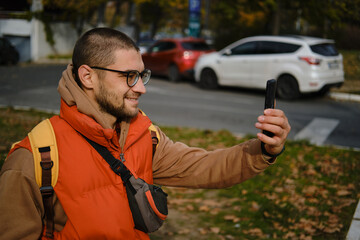 Smiling man holding smartphone during video call in autumn city park. Travel blogger concept