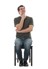 Thoughtful young man seated on a chair keeps hand under chin looking joyful aside, thinking positive. Contented male sitting isolated on transparent background